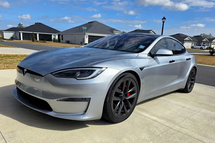 Silver electric sedan parked in a residential driveway under a blue sky with scattered clouds.