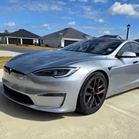 Silver electric sedan parked in a residential driveway under a blue sky with scattered clouds.