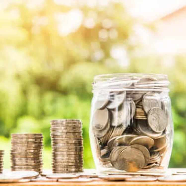 Stacks of coins and a glass jar filled with coins sit on a wooden table.