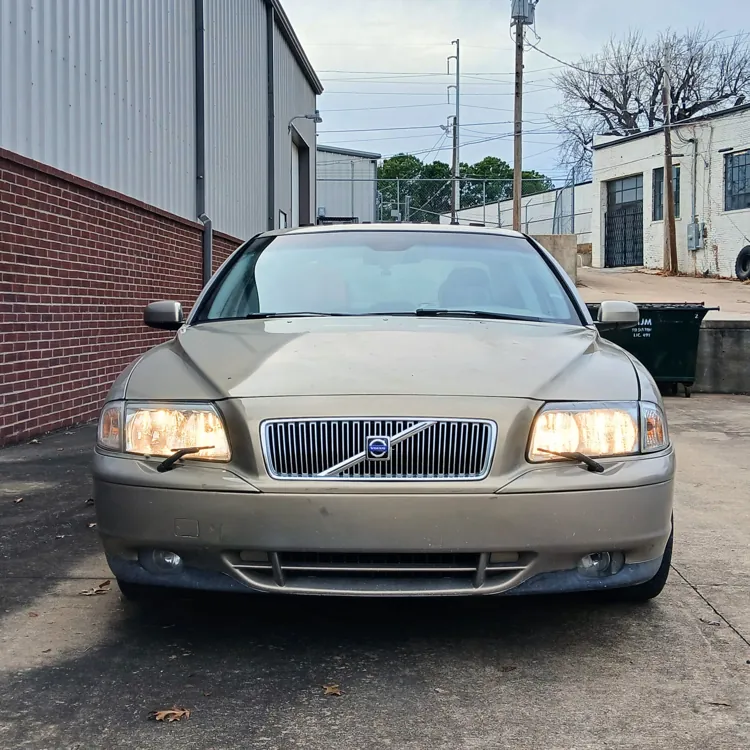 Front view of a beige sedan parked in an industrial area with brick and metal buildings.