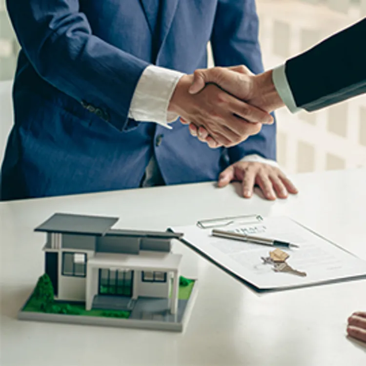 two men shaking hands with a model house and paperwork on a desk